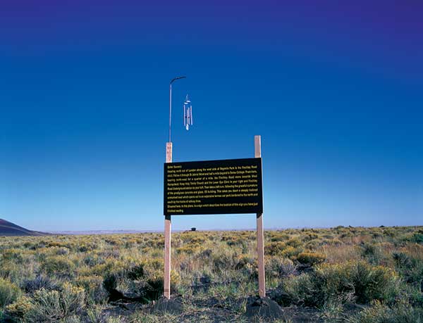 “Better Scenery, 2000,” by Adam Chodzko, presents language in the landscape, but it refers to elsewhere. Here in the Painted Desert, the reader finds directions from central London to another sign in the O2 shopping center car park. The wind chime atop the sign alerts the reader’s body to here.