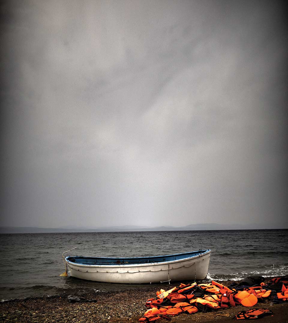 Orange lifejackets piled up on the shore of an island just next to a boat moored nearby. The open ocean and a dark sky looms in the background.