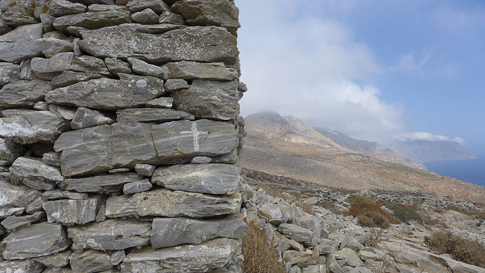 A small cross endures on one stone inside of a wall that overlooks a rocky mountain plain.