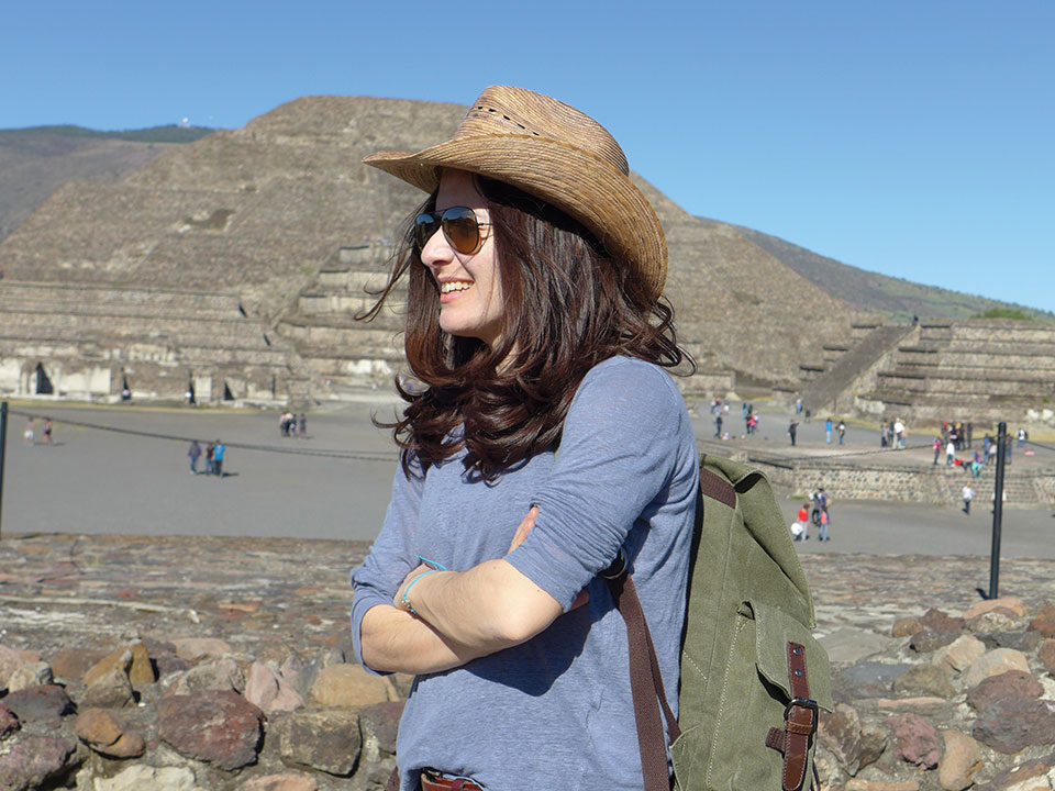 Eleni Kefala stands, arms crossed and smiling, in profile, wearing a straw hat