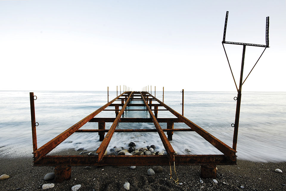 The steel frame of a dock, not covered and thus exposing the sea below, extends out from a rocky beach.