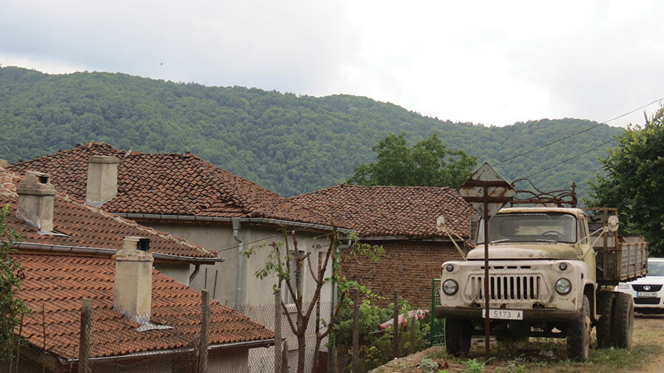 A large truck sits on the road just above a village with forested mountains in the background
