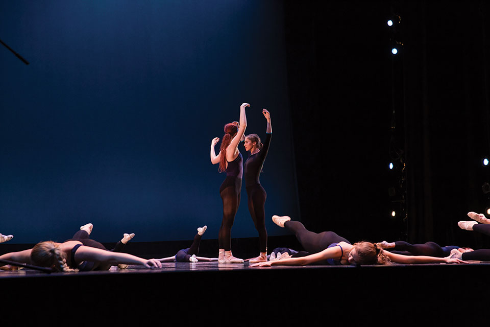 Two dancers stand face to face standing amidst other dancers lying motionless on the stage
