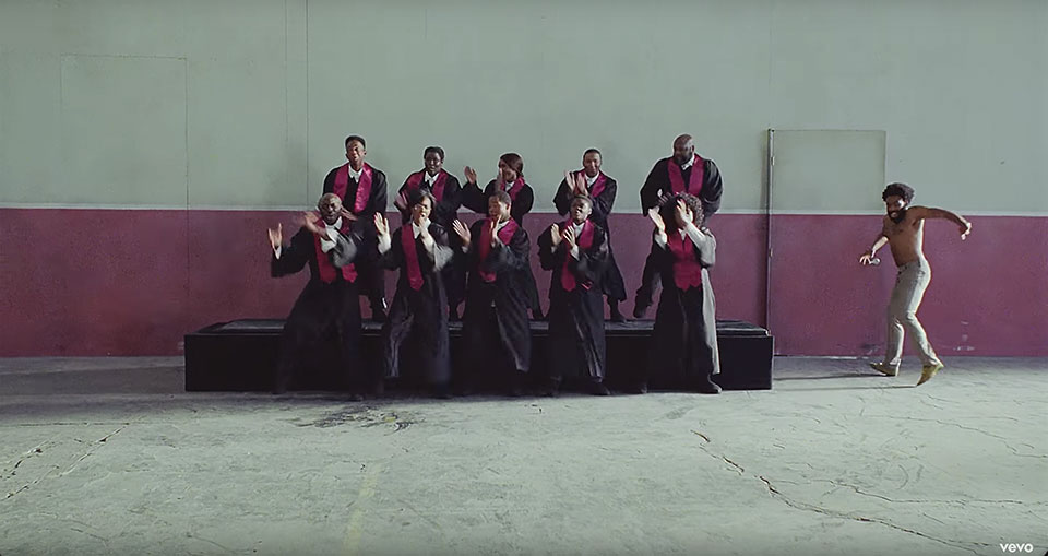 A still image from the video to Childish Gambino's "This is America," featuring a choir of African-Americans singing on riser as Gambino emerges into the frame from the right