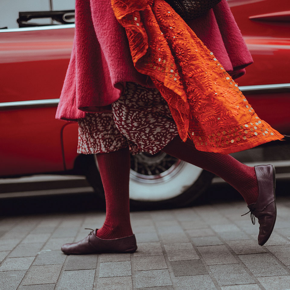 The lower half of a women, dressed mostly in reds and oranges, walking along a busy city street