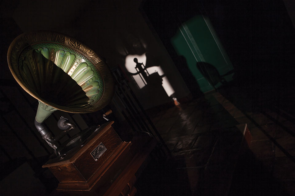 A Victrola, partially draped in shadow, in the foreground as fading light is swallowed up by a room tilted askew in the background