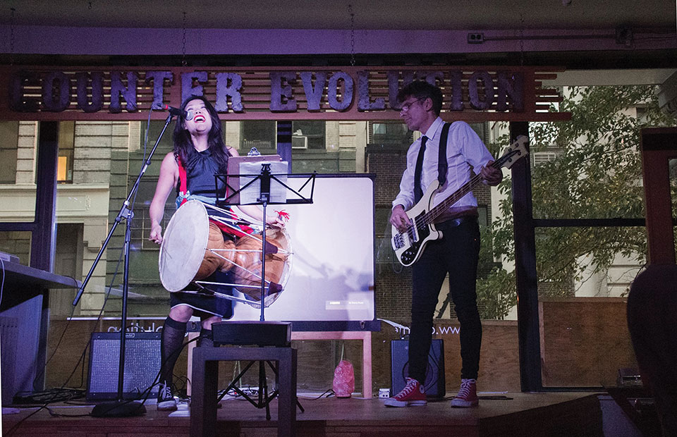 Two musicians, a woman playing a bass drum and a man playing a bass guitar, perform on stage