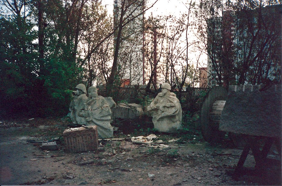 A collection of statues of resting soldiers, tucked into a small grove of trees with trash and industrial detritus strewn about