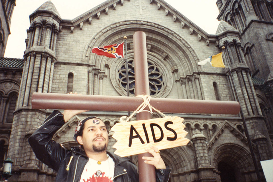 A man in a leather jacket carries a cross with a sign hanging around it that reads, "AIDS," while standing in front of a church