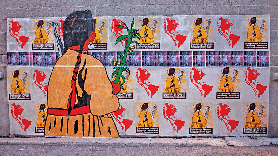 A wall covered in an image dominated by a large human figure that appears to be looking at a recurring pattern of that same figure looking at an outline image of North and South America
