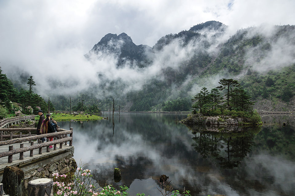 Spectators stand on a stone overlook that juts on to a wooded lake that sits at the foot of foggy mountains