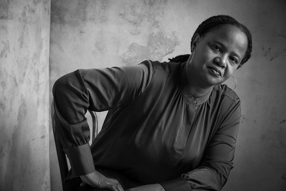 A black and white photo of Edwidge Danticat, seated but leaning forward slightly, with her hand on her leg