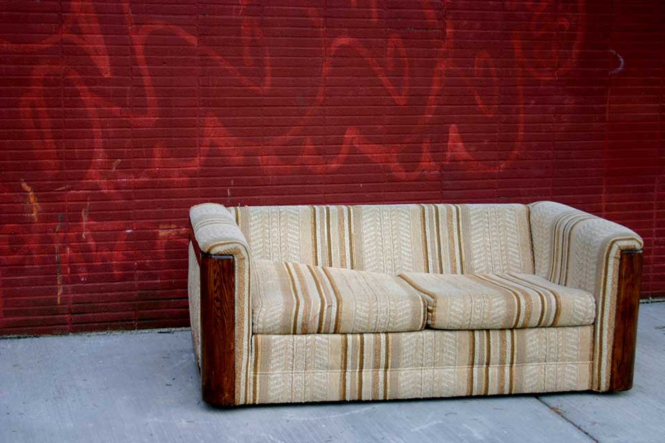 A photograph of a sofa sitting in front of a wall, painted red with faded graffiti still visible