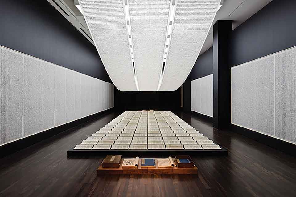 A photograph of an art installation. A long table sits beneath a long, curved light. The table is covered with carefully arranged parchment with writing on them.