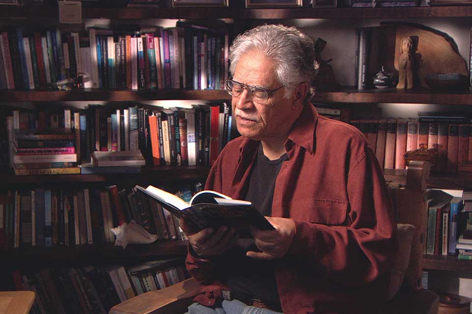 A photograph of Rudollfo Anaya reading a book in his study