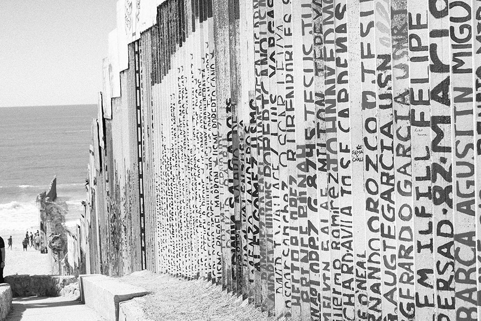 A black and white photograph of a fence made of upright slats, each with words written on them vertically