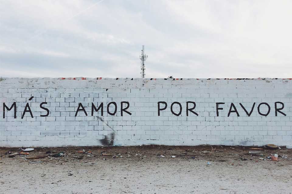 A photograph of a long wall with graffiti on. Text reads: Mas Amor Por Favor 