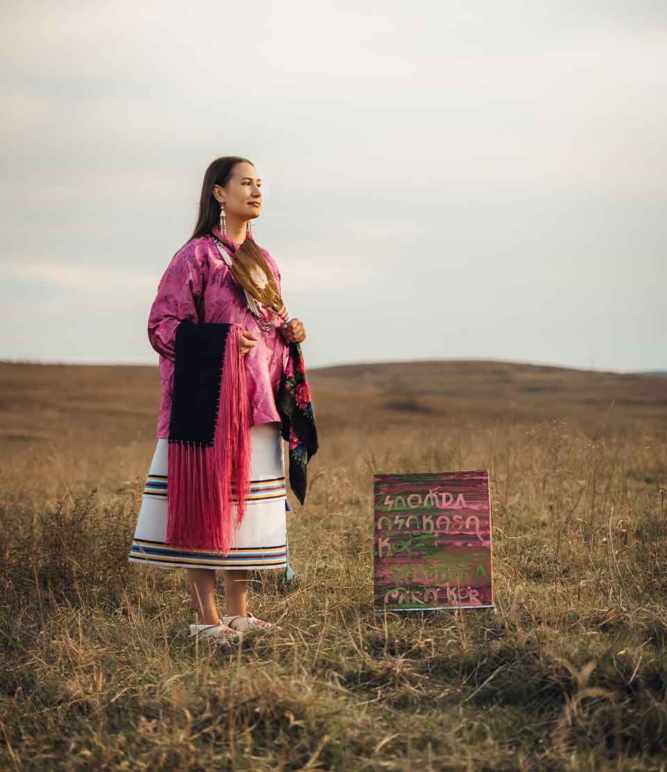 A photograph of a woman in indigenous garb standing on scrub grass hilltop with a painting nearby
