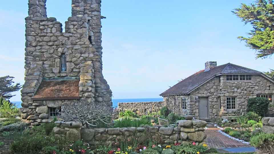 A rugged tower rises to the left of a stone house, both of which sit at the edge of the ocean