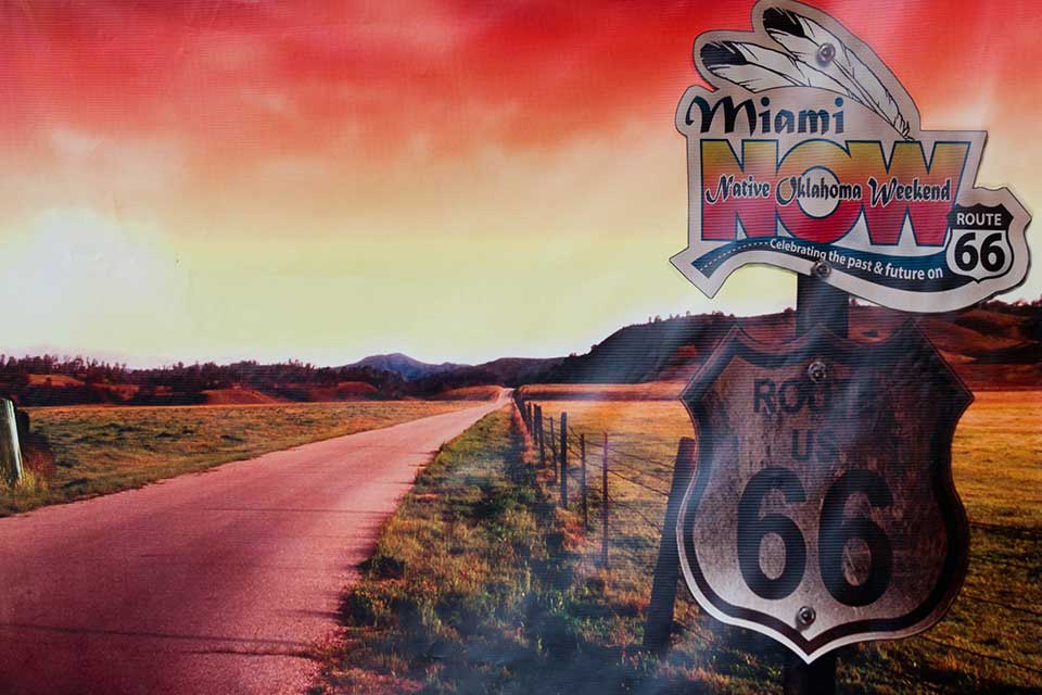 A color-altered photograph (emphasizing red tones) of a highway with a sign beside it in the foreground. The sign reads Route US 66. Another sign has been placed above it that read Miami Now, Native Oklahoma Weekend. Celebrating the past and future on Route 66.