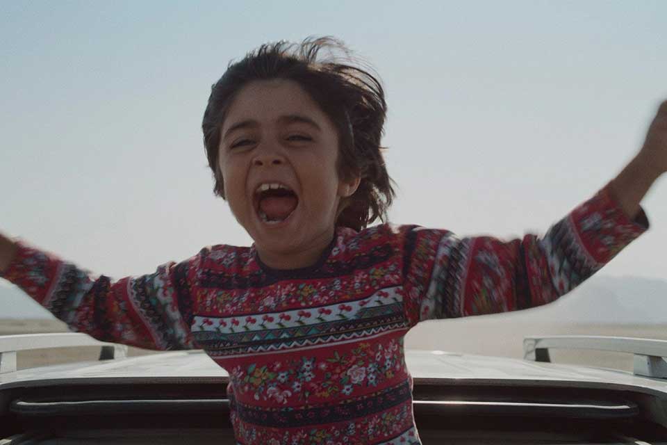 A still image of a boy with his arms outstretched coming out of the top of a moving car