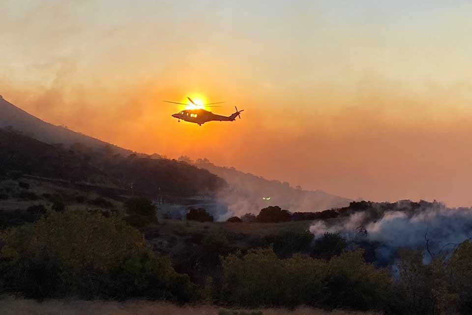 A helicopter hovers above a smoldering, mountainous landscape. The sun is positioned just such that the helicopter appears to be on fire.