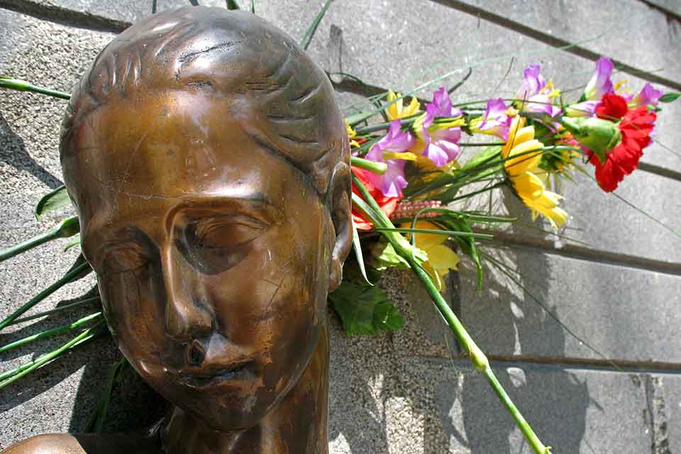 A photograph of a the head of a bronze statue of a female figure. Flowers have been placed behind the statue's head