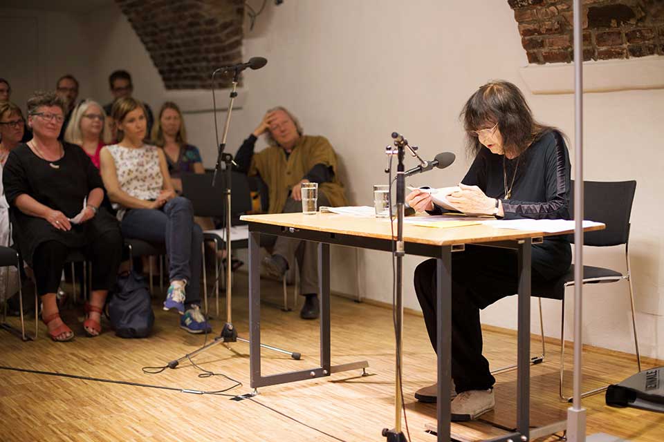 Friederike Mayröcker sits at a table reading with a small group sitting to the side