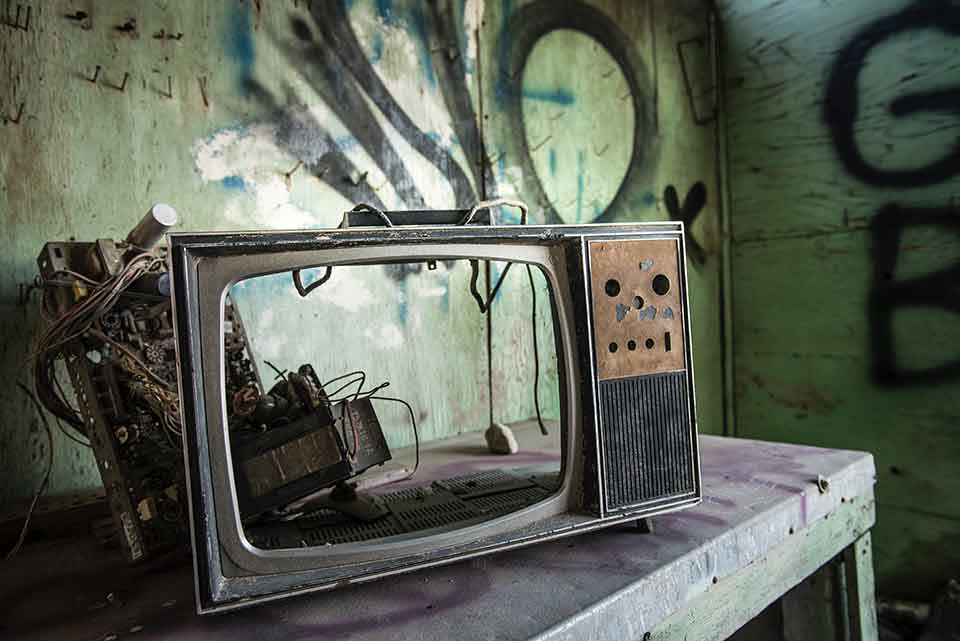 A photograph of a broken television with no screen in a room tagged with graffiti on the walls
