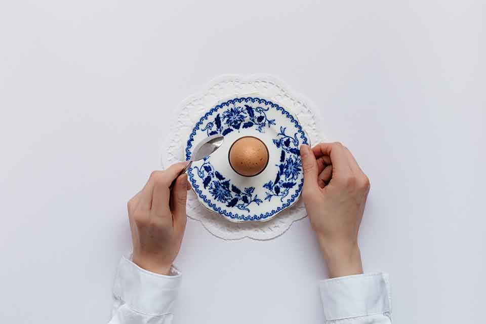 A photo looking down on a single egg on a fine piece of blue china, framed by two hands preparing to eat the egg