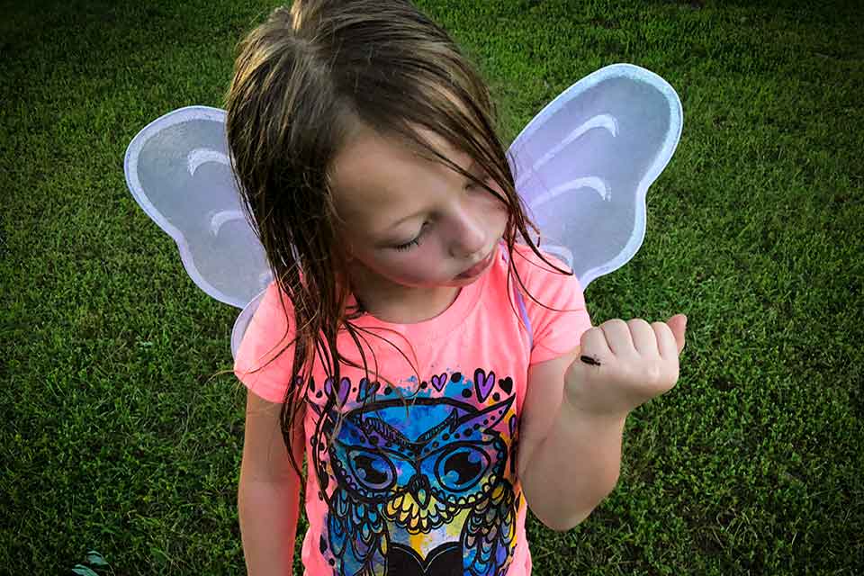 A photograph of a young girl with wet hair in a pink t-shirt. She is wearing a pair of translucent wings.