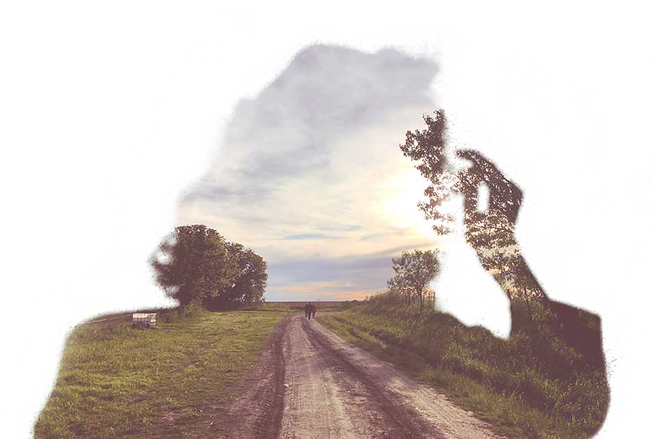 The negative outline of a person blowing on a dandelion through which a photograph of a dirt road stretching into the distance can be seen