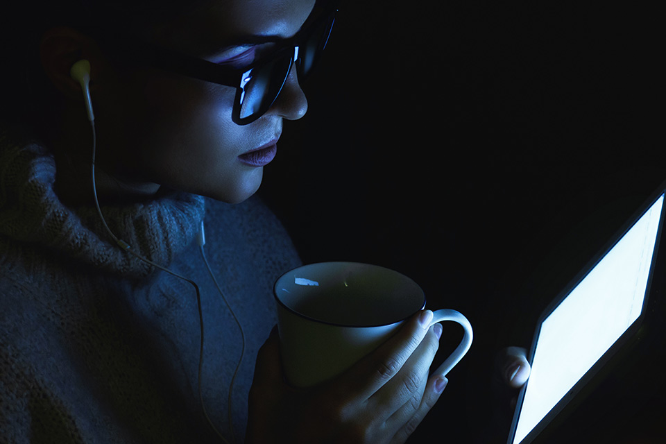 A woman drinking a cup of coffee, bathed in blue light from a computer screen.