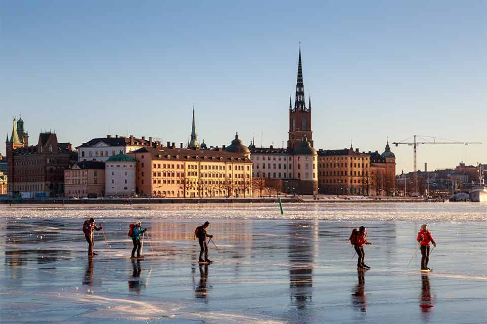 A photograph of skaters on a frozen lake with old city buildings in the background