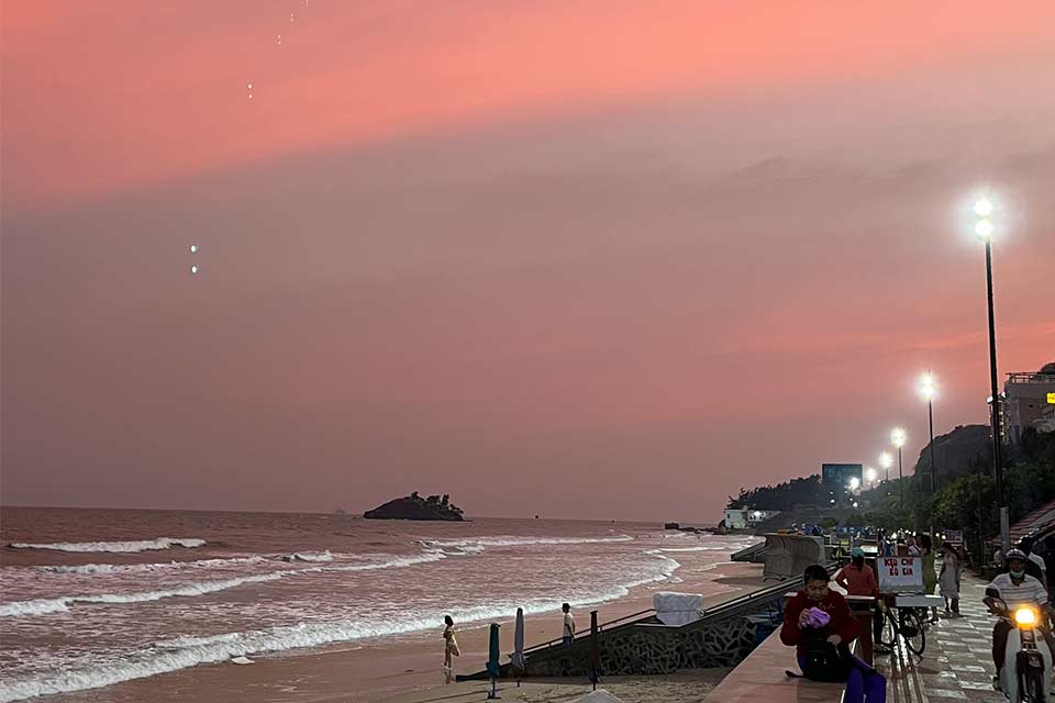 A photograph of a beach at sunset with a lighted walkway full of people running beside it