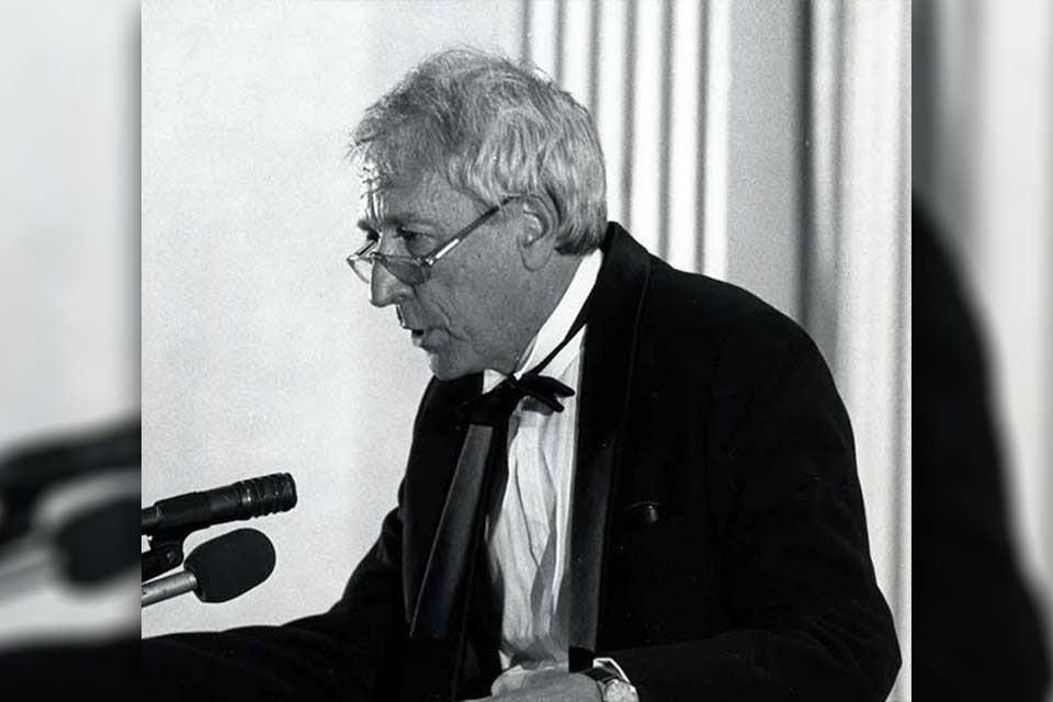 A black and white photograph of Tomas Transtromer talking into a microphone at a lectern.