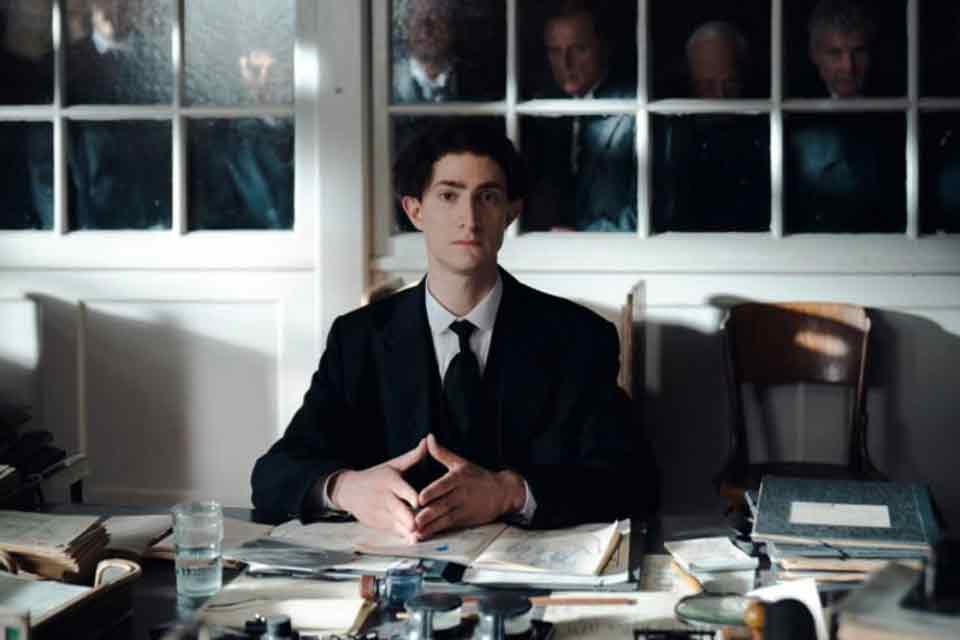 A man in a suit sits at a cluttered desk, looking directly at the viewer