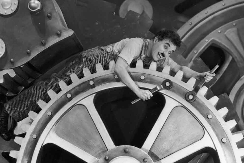A man with a wild smile riding a large gear as he works to repair large nuts on the gear with twin wrenches