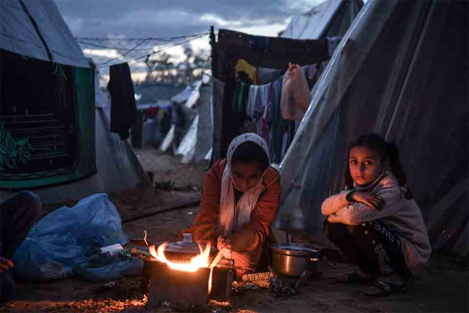 Three figures sit around a small fire outside of a canvas tent