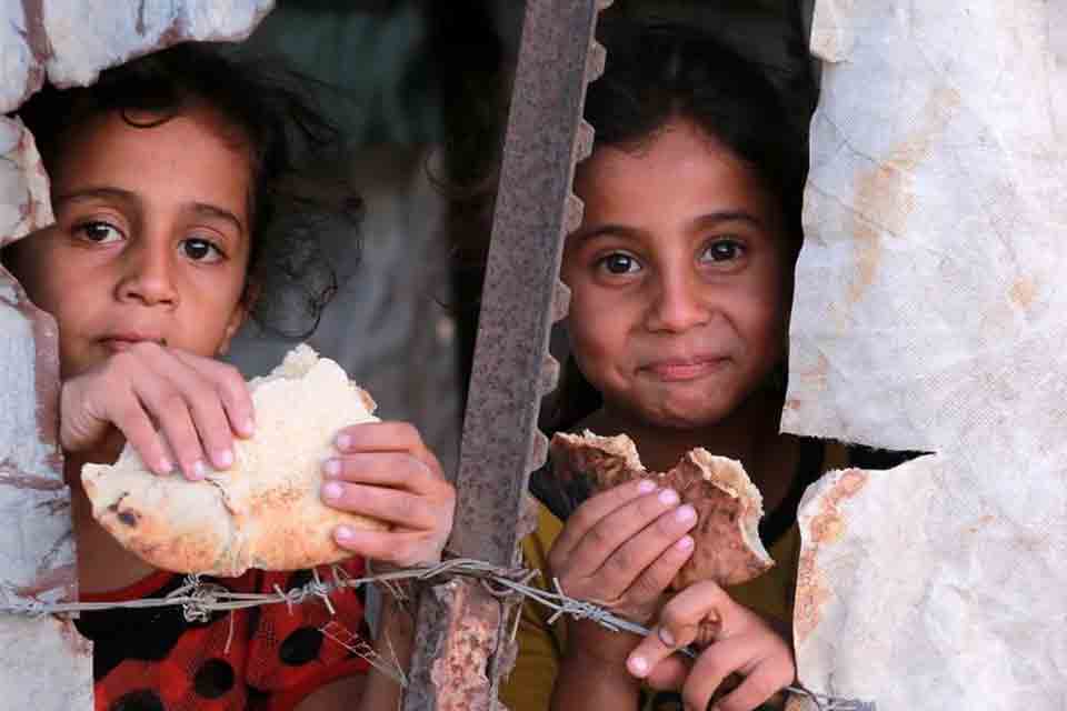 Two children eating bread peer through a crack in a ruined structure