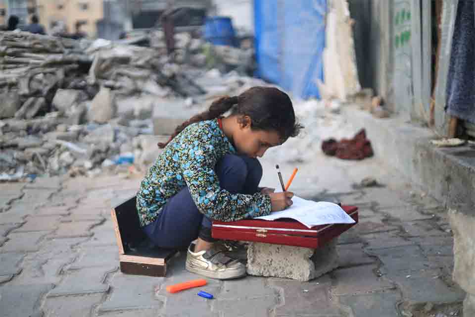 A young girl kneels in the rubble to use a displaced stone as a desk to work on schoolwork