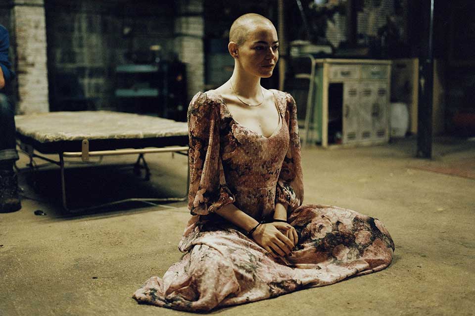 A bald woman sitting calmly in an ornate dress on a grubby floor