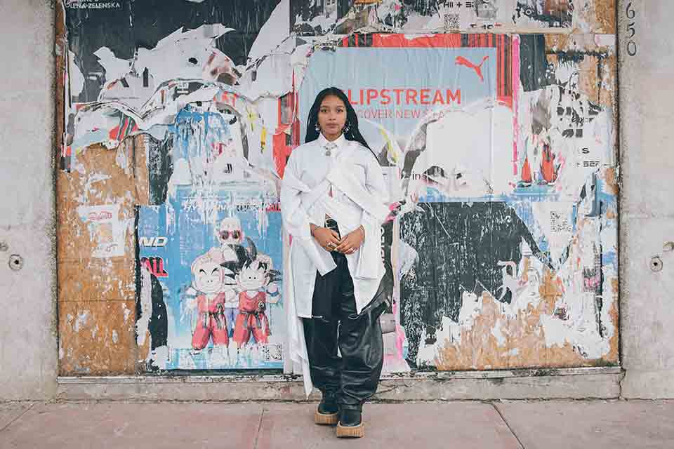 A photograph of a dark skinned woman standing in front of a urban wall