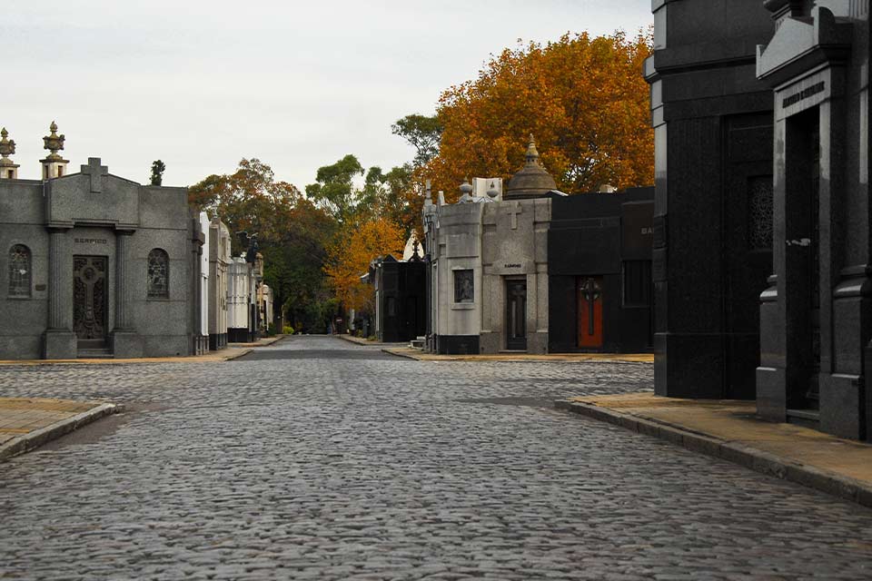 A photograph of a small-stone paved road, lined by ornate mausoleums, inside a tidy cemetary
