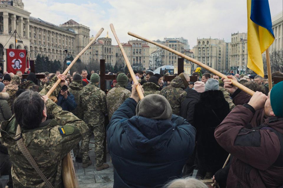 A photograph of a group of three people holding long wooden poles to their face in a crowd of protesters