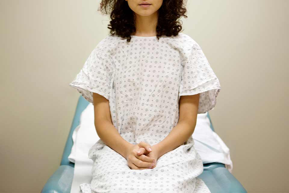 A woman in a hospital gown sitting on an examination table with her hands folded in her lap