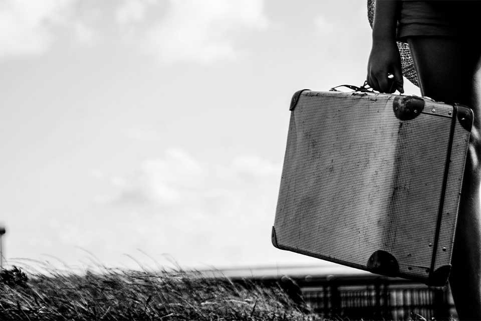 A black and white photograph of a hand attached to a figure in a business suit, holding a weathered suitcase