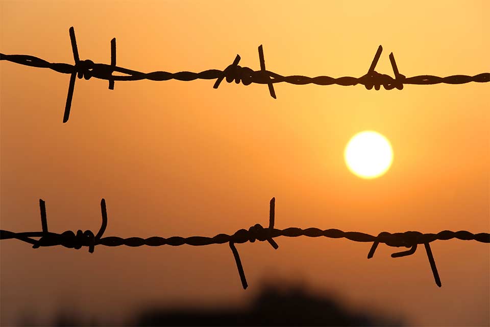 A bleary orange sun is setting, as seen through a barbed wire fence.