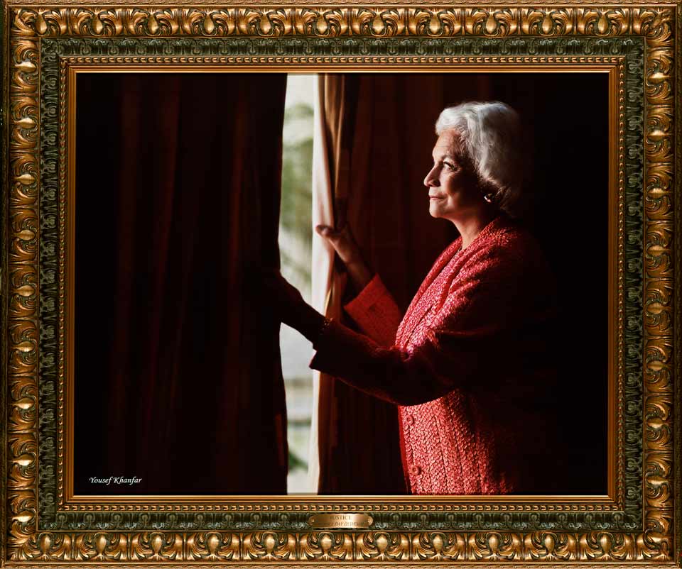 A photograph of Justice Sandra Day O'Connor peering out through a heavy curtain to the daylight outside