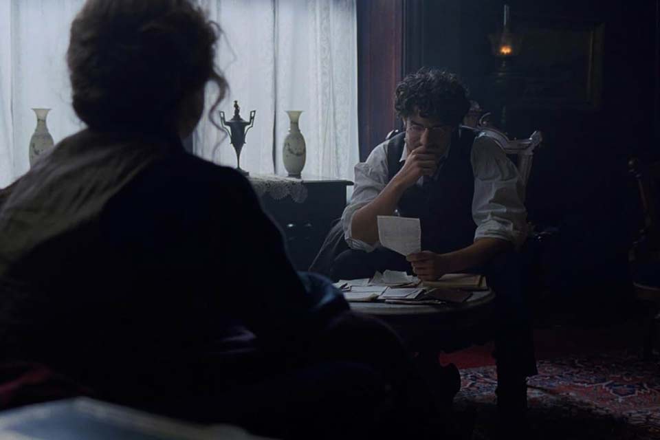 A figure with their back to the viewer looks on as a seated man bemusedly looks at a piece of paper